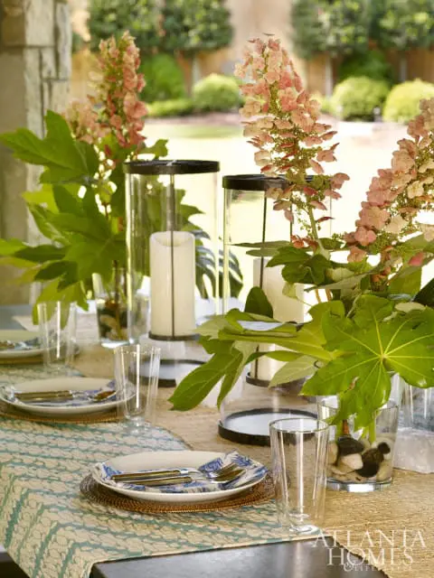 Outdoor dining table set with blue and white patterned plates, glassware, and floral centerpieces in hurricane lanterns.