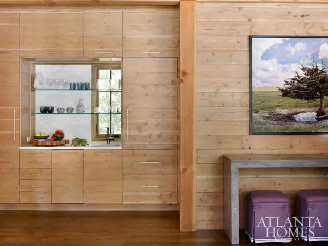 Light wood paneled walls and cabinetry with brass hardware, marble backsplash, glass shelves displaying glassware and decorative bowls, and a sink with a window view.