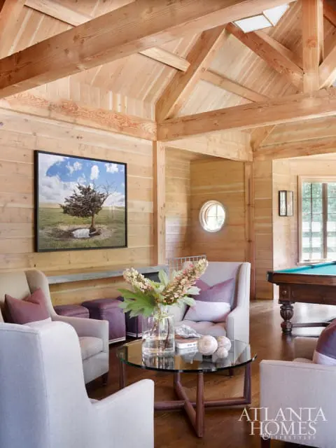 Living room with wood-paneled walls and vaulted ceiling, featuring plush seating, a glass coffee table with floral arrangement, and a pool table in the background.
