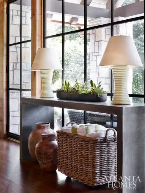 Console table with two ribbed ceramic lamps, a planter of succulents, two terracotta vases, and a wicker basket filled with towels. Large windows with stone accents are visible in the background.