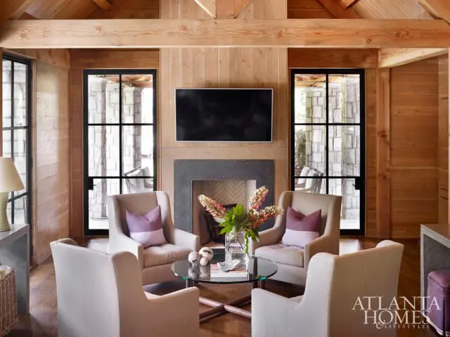 Living room with wood-paneled walls and ceiling, stone fireplace, four upholstered armchairs, and a glass coffee table.