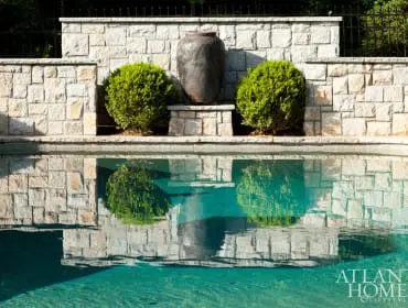 Stone retaining wall with two rounded bushes and a large dark urn, reflected in a swimming pool.