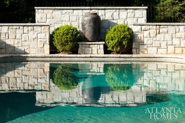 Stone retaining wall with two rounded bushes and a large dark urn, reflected in a swimming pool.