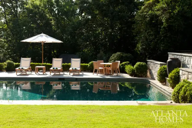 Outdoor swimming pool with lounge chairs, a table and chairs, and lush landscaping. A large dark urn sits on a stone wall.