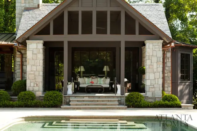 Covered porch with stone pillars and large windows, furnished with a sofa and coffee table, overlooking a swimming pool.