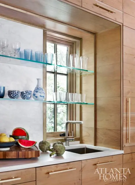 Wood cabinetry with brass hardware, marble countertops, and glass shelves displaying glassware and blue and white ceramic dishes. A stainless steel faucet is over a sink, with a window behind.
