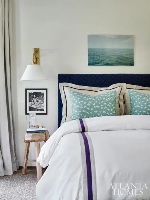 Bedroom with white shiplap walls, navy upholstered headboard, white duvet with purple and gray stripes, and coastal artwork above the bed.