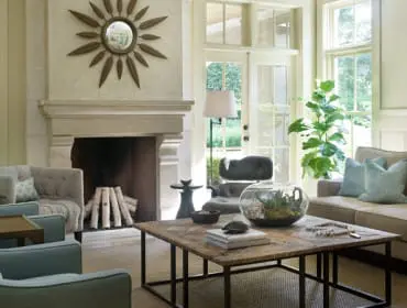 Living room with a large stone fireplace, tufted gray armchair, light brown sofa with blue pillows, and a rustic wood coffee table.