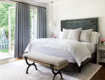 Bedroom with white shiplap walls, a grey upholstered headboard, white bedding, and grey curtains opening to a garden view.
