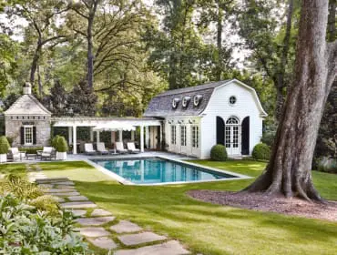 White pool house with arched windows and dormers next to a rectangular pool. Stone cabana with pergola and lounge chairs in a lush, green garden with a swing hanging from a large tree.