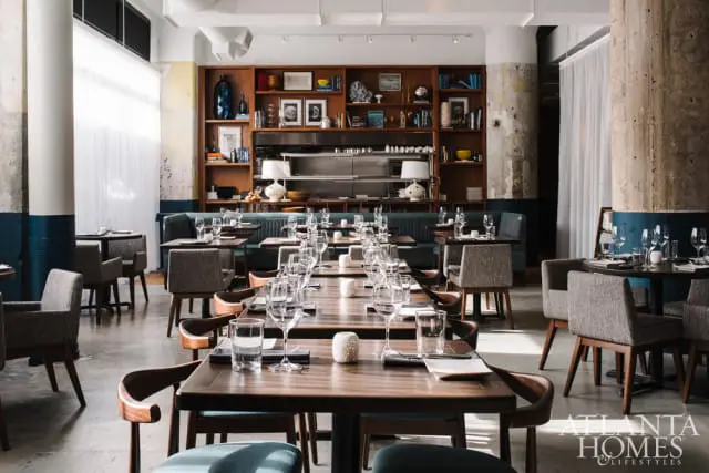Restaurant dining room with dark wood tables set with glassware and place settings, upholstered chairs, and a large wooden bookshelf filled with books and decor.