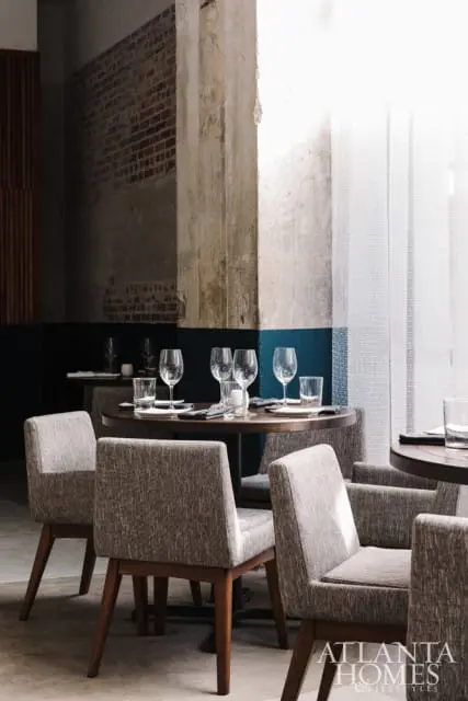 Restaurant dining room with round tables set for dinner, upholstered chairs, exposed brick walls, and concrete columns.