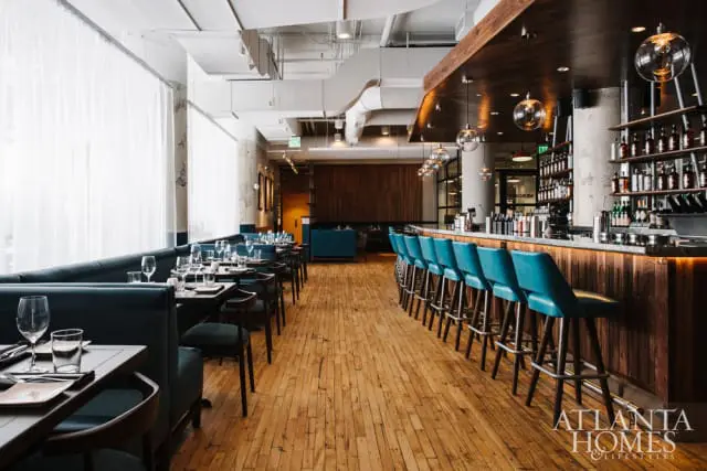Restaurant interior with teal banquettes and bar stools, wood floors, and a long wooden bar stocked with bottles.