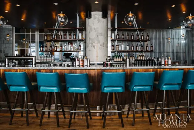 Bar with teal leather stools, marble countertop, and shelves stocked with liquor bottles. A stained glass window is visible behind the bar.