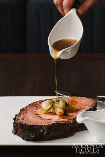 A slice of prime rib is topped with onions and herbs as a hand pours gravy from a white pitcher.