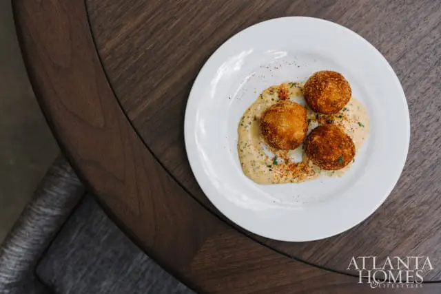 Three golden-brown fried arancini balls served on a white plate with creamy sauce and sprinkled spices, on a dark wood table.