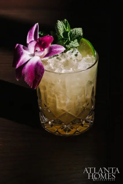 Crystal rocks glass filled with ice and a light-colored cocktail, garnished with a purple orchid, mint sprig, and lime wedge.