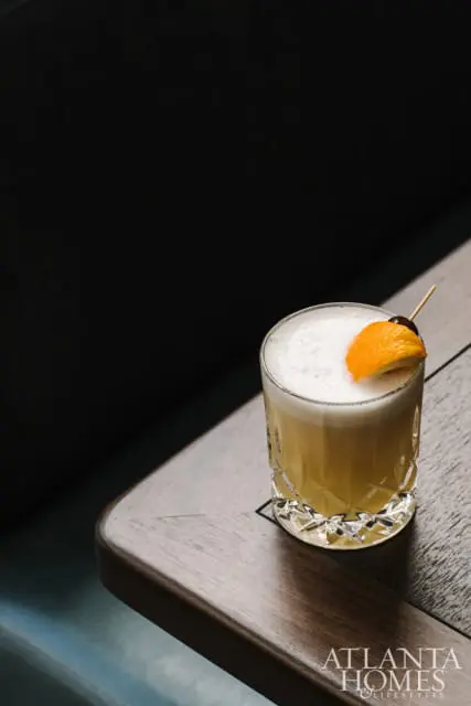 A cocktail in a cut glass tumbler with a frothy top, garnished with an orange slice and cherry, sits on a dark wood table.