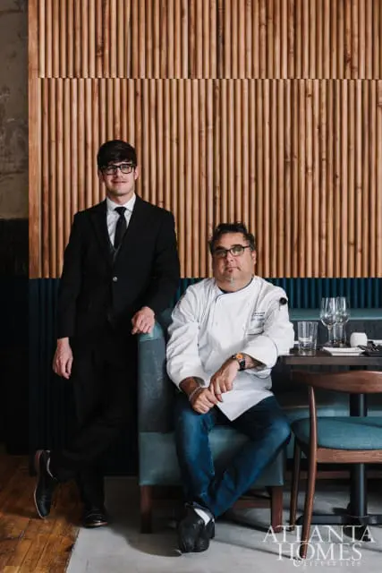 A man in a suit and glasses stands next to a chef in a white coat and jeans, seated in a restaurant booth with a wood-paneled wall behind them.