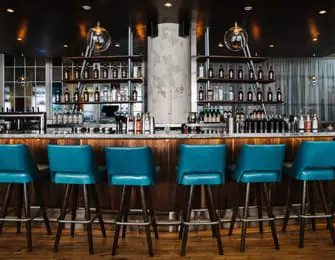 Bar area with teal upholstered stools, wood paneling, marble countertop, and shelves stocked with bottles and glassware.