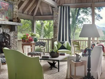 Screened porch with stone fireplace, light green slipcovered furniture, patterned chairs, and mountain views through large windows