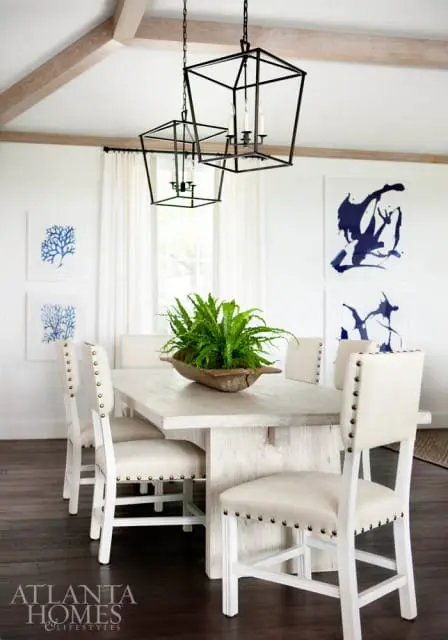 Dining room with a light wood table, upholstered chairs, black lantern pendant lights, and abstract blue art on white walls.