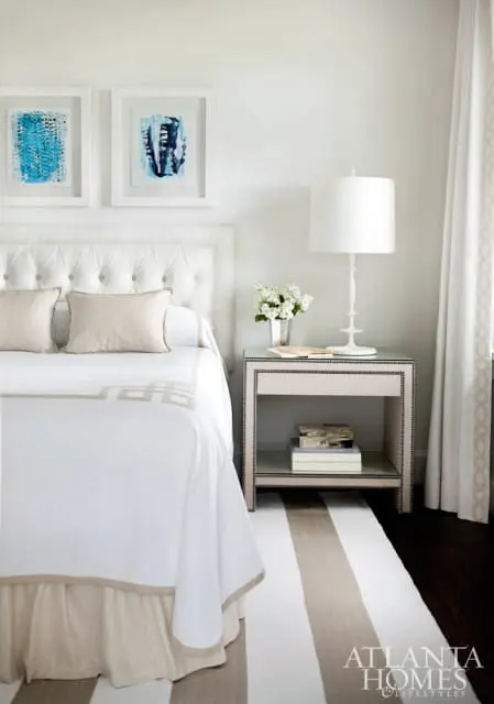 White tufted headboard with beige pillows on a bed with white bedding and a beige skirt. A bedside table with a white lamp and a vase of flowers sits next to the bed.