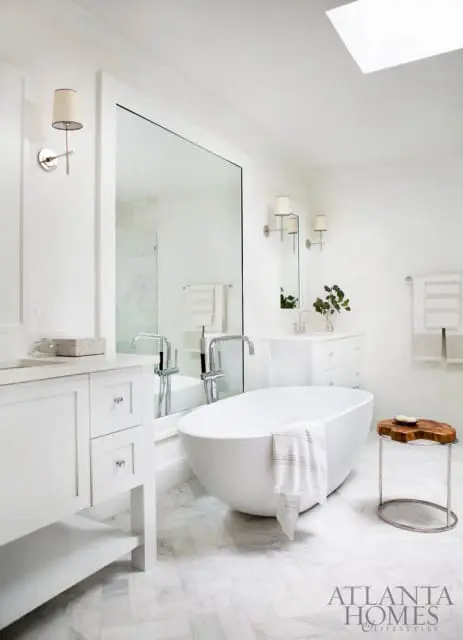 White bathroom with freestanding soaking tub, marble herringbone floor, double vanity, and large mirror reflecting a glass-enclosed shower.