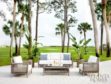 Outdoor living area with white sofa, chairs, and coffee table on a patio, surrounded by palm trees and a green lawn overlooking the ocean.