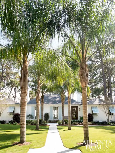 White brick house with light blue shutters and a dark wood front door, framed by palm trees and a manicured lawn.