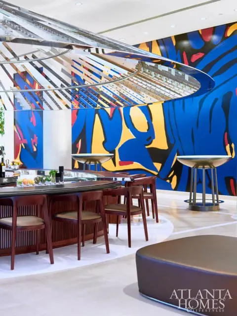 Modern bar area with a curved wooden bar, leather stools, and a vibrant abstract mural on the wall. A metallic, circular light fixture hangs overhead.