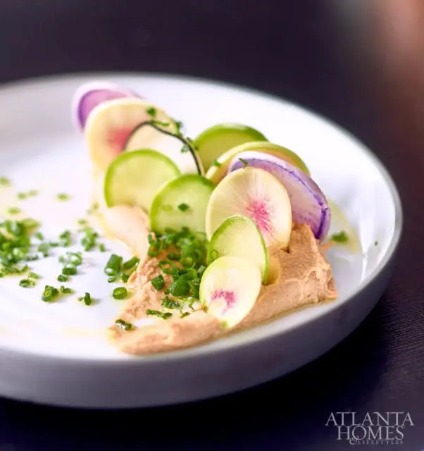 Close-up of a white plate with a dollop of hummus topped with thinly sliced radishes and cucumbers, garnished with chopped chives.
