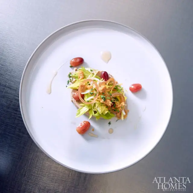 A plate of food with sliced meat, celery, red grapes, and crispy onions, drizzled with sauce. The Atlanta Homes & Lifestyles logo is in the bottom right corner.