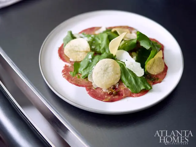 Beef carpaccio with arugula, parmesan shavings, and potato crisps on a white plate, with Atlanta Homes & Lifestyles logo in the corner.