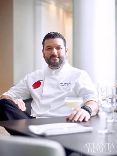 Julio Delgado, Executive Chef, wearing a white chef's coat with a red and blue emblem, sits at a table with a cocktail and place setting.
