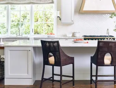 Light gray kitchen with white marble countertops, herringbone tile backsplash, and dark woven bar stools. A large window overlooks greenery.