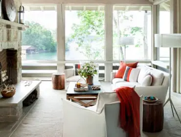 Living room with stone fireplace, white sofas, and large windows overlooking a lake. Orange throw pillows and blanket add pops of color.