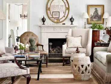 Living room with fireplace, ornate mirror, upholstered chairs, and a tufted leather armchair. A bust sits on a coffee table with books.