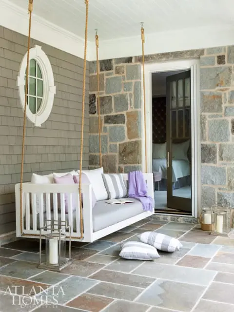 White porch swing with gray and white striped cushions and pillows, purple throw, and open book. Stone wall and slate tile floor.