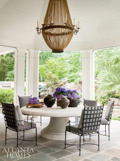 Covered patio with a large round stone table and chairs, a rope chandelier, and purple hydrangeas. The Atlanta Homes & Lifestyles logo is in the lower left corner.
