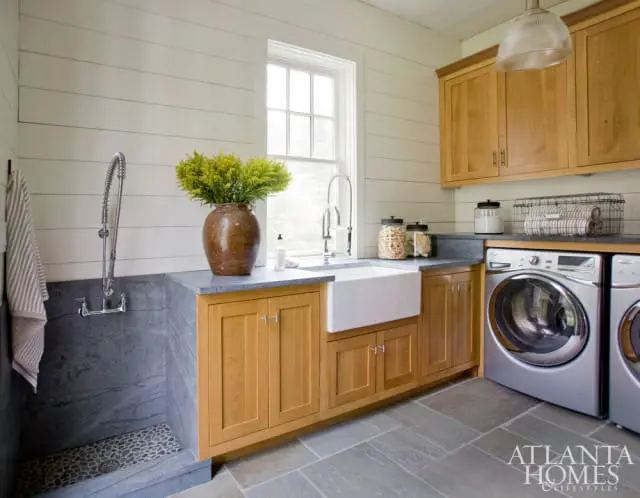 Laundry room with white shiplap walls, a large white farmhouse sink, wooden cabinetry, a dog washing station with stone flooring, and stainless steel washer and dryer.