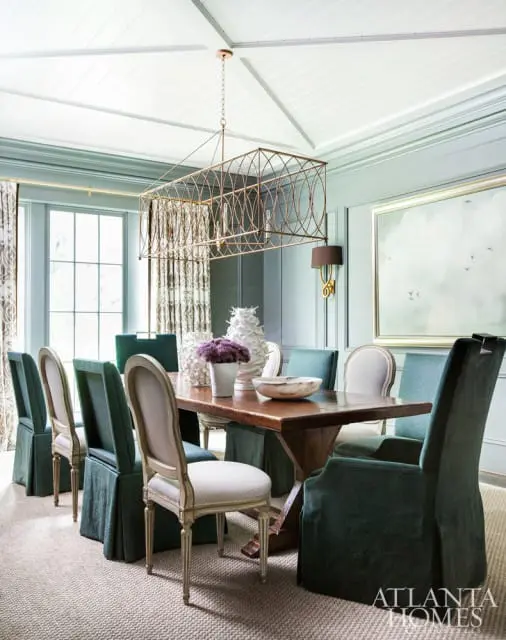 Dining room with a rectangular wooden table surrounded by upholstered chairs in teal and cream. A large gold geometric chandelier hangs from the vaulted ceiling.