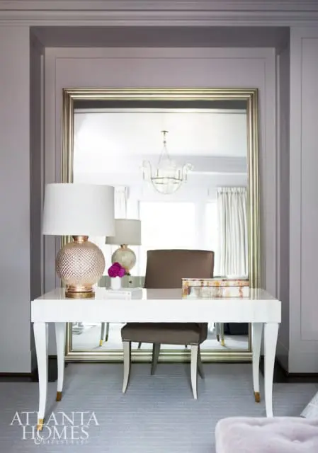 White desk with a textured rose gold lamp, books, and a decorative box. A brown upholstered chair sits behind the desk, reflected in a large gold-framed mirror.