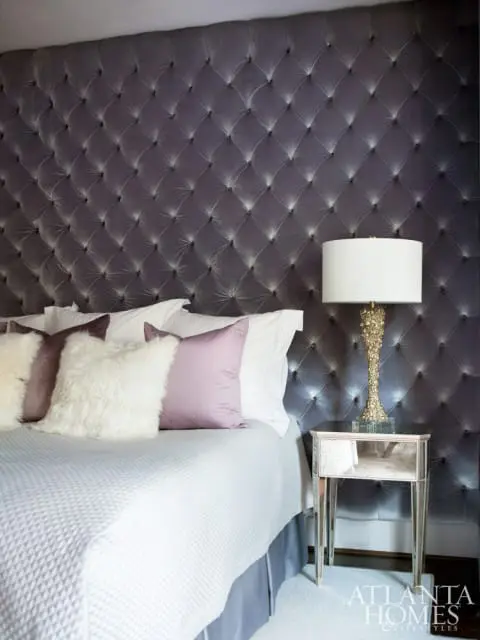 Bedroom with a large, dark gray tufted headboard, white bedding, plush pillows, and a mirrored nightstand with a decorative lamp.