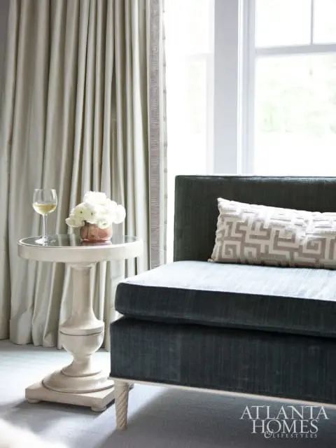 Dark teal sofa with a geometric pattern lumbar pillow next to a pedestal side table with a glass top, a wine glass, and a vase of white flowers.