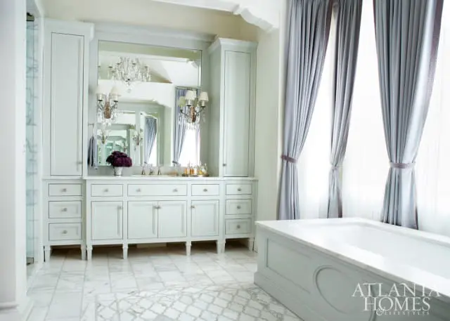 Light blue bathroom vanity with marble countertop and floor, flanked by tall cabinets. A large mirror reflects a chandelier and sconces. A bathtub sits to the right, with grey curtains framing the window.