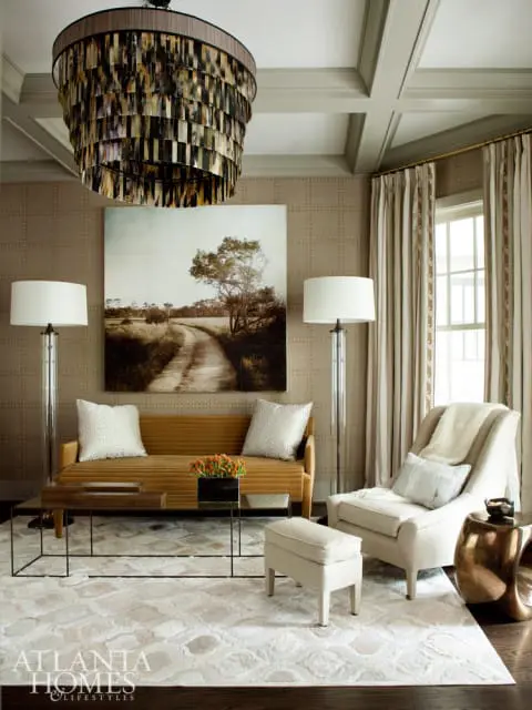 Living room with a textured tan wallpaper, coffered ceiling, and a large tiered chandelier. A mustard-yellow sofa faces a coffee table and a cream armchair.