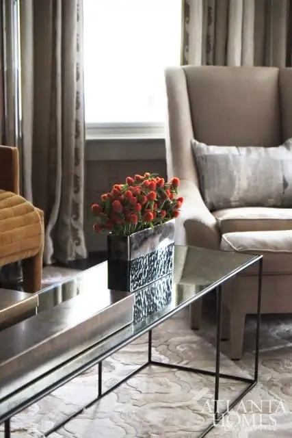 Living room with a beige wingback chair, a mirrored coffee table with a vase of orange flowers, and patterned curtains.