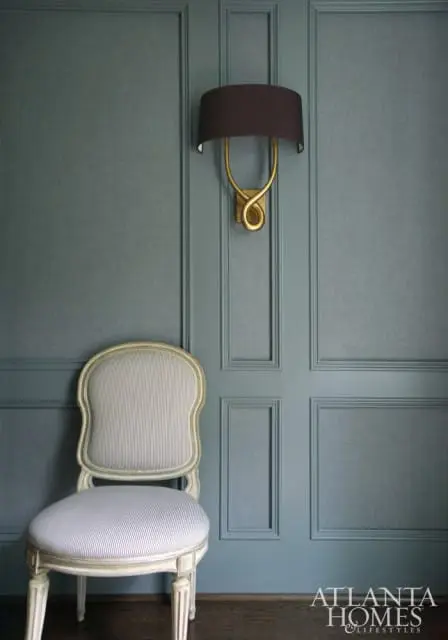 Blue paneled wall with a gold sconce and a striped upholstered chair with a distressed wood frame
