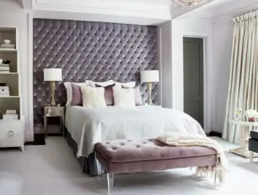 Bedroom with a large tufted purple headboard, white bedding, a purple bench, and a white bookshelf filled with books and decorative items.
